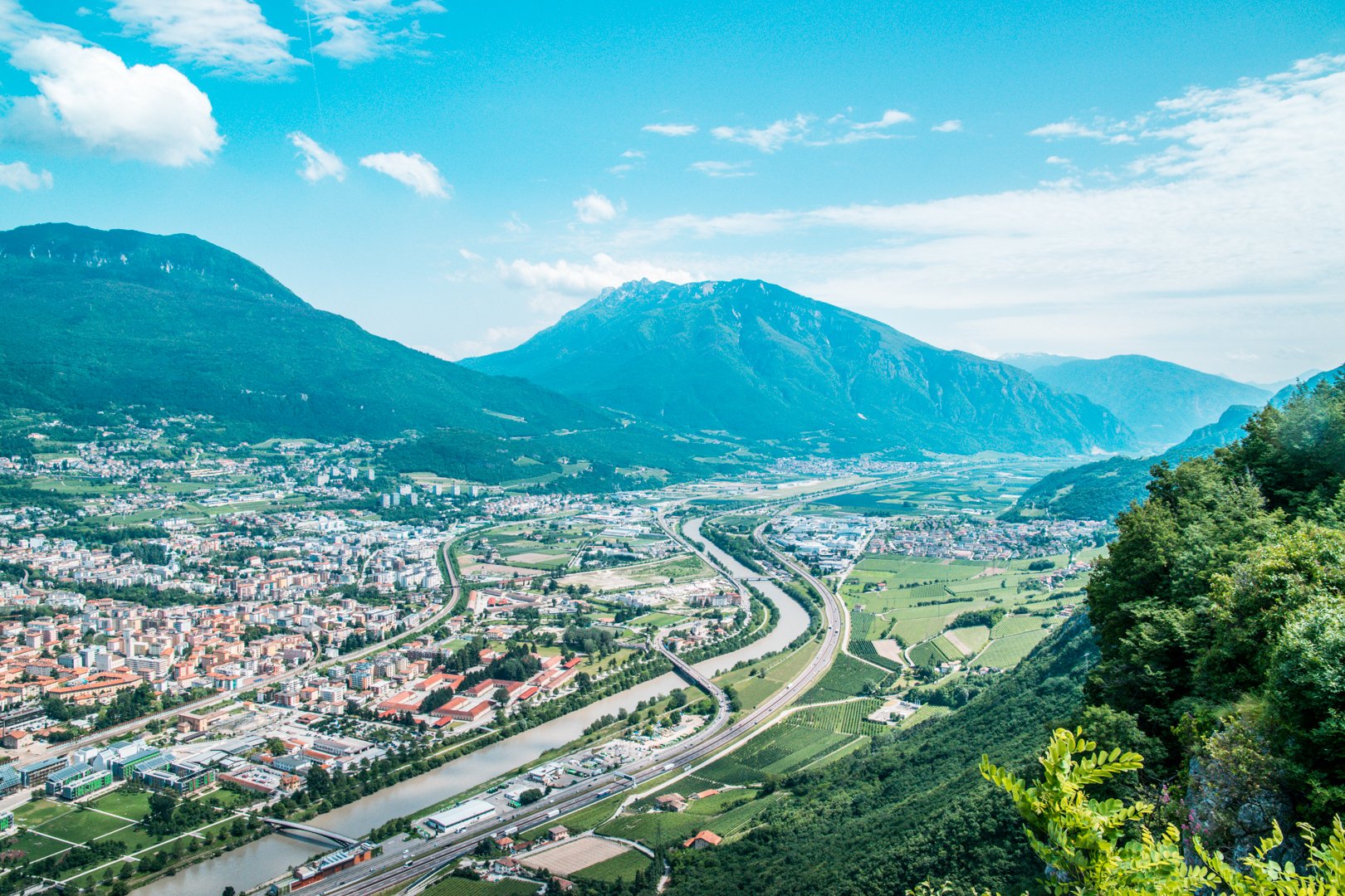 Wederom een uitzicht over Trento vanaf het panorama-terras boven op de berg. Je ziet de Aldige-rivier door het landschap kronkelen, en op de achtergrond bergen die zo hoog zijn dat ze tot de wolken rijken. Links in de hoek zie je nog een deel van Trento. 